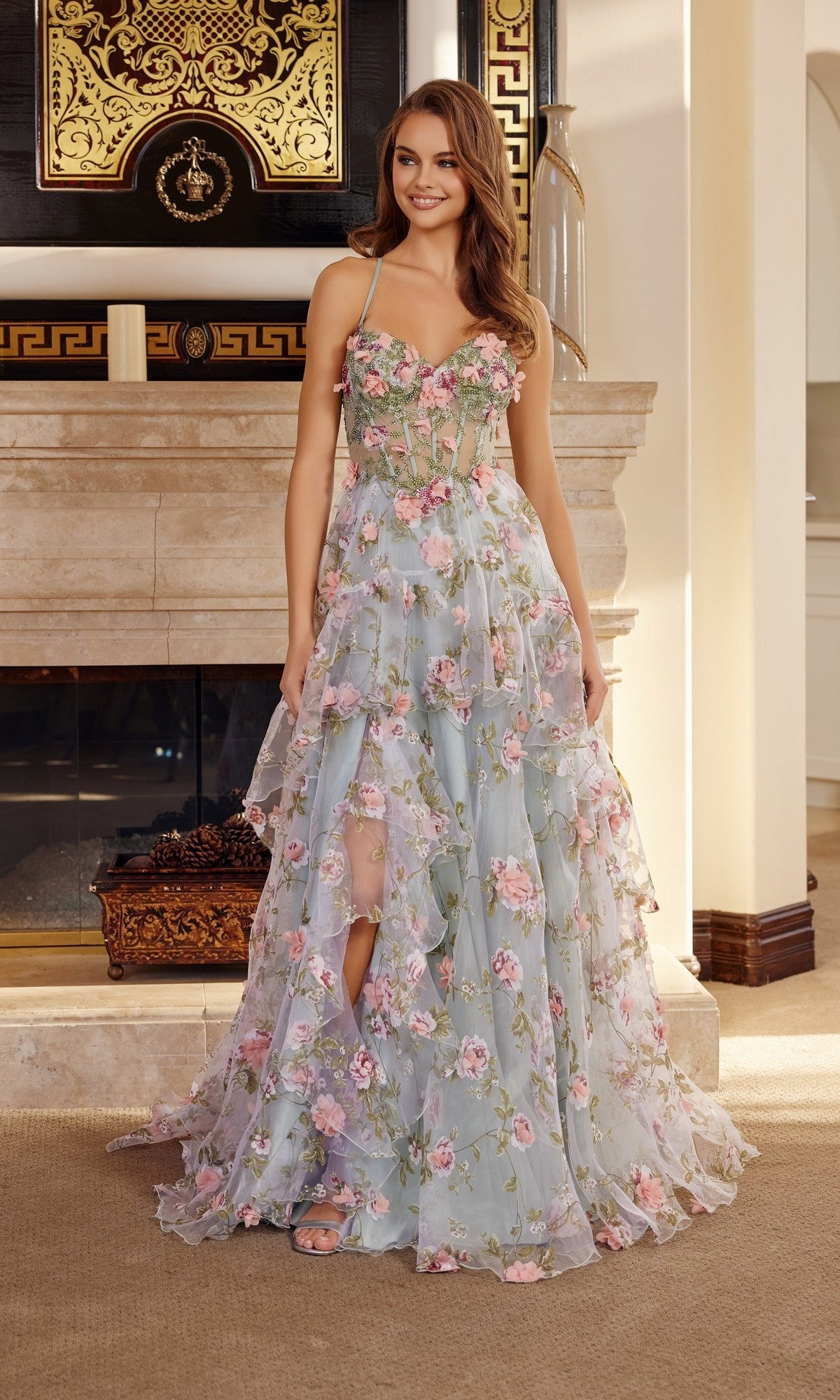 A woman stands indoors, smiling and posing in front of a fireplace while wearing the Nox Anabel G1783 long green prom dress, featuring 3D floral appliques and tiered ruffles.